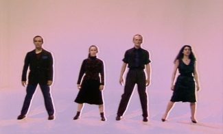 Four people stand in a line against a light-colored background, posing in coordinated dance stances.
