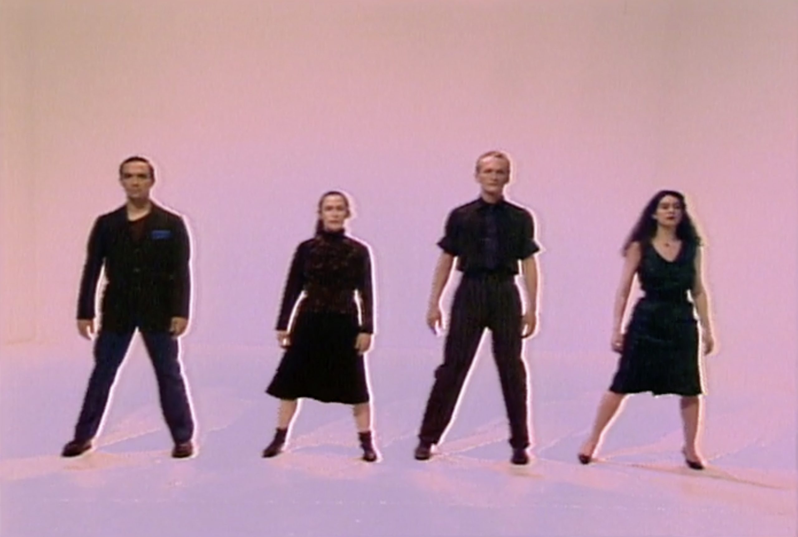 Four people stand in a line against a light-colored background, posing in coordinated dance stances.