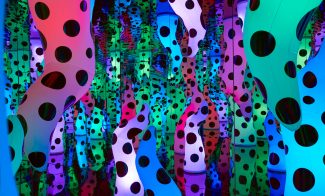 A mirrored room filled with illuminated, organic-shaped sculptures covered in large black polka dots, glowing in shades of pink, blue, and green, as seen in Love is Calling at the ICA Boston.