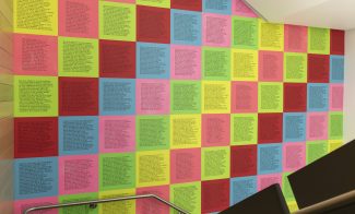 A wall covered in a grid of colorful square posters, each containing columns of printed black text, displayed beside a staircase with a metal handrail.