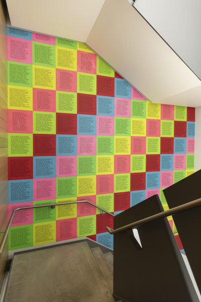 A wall covered in a grid of colorful square posters, each containing columns of printed black text, displayed beside a staircase with a metal handrail.